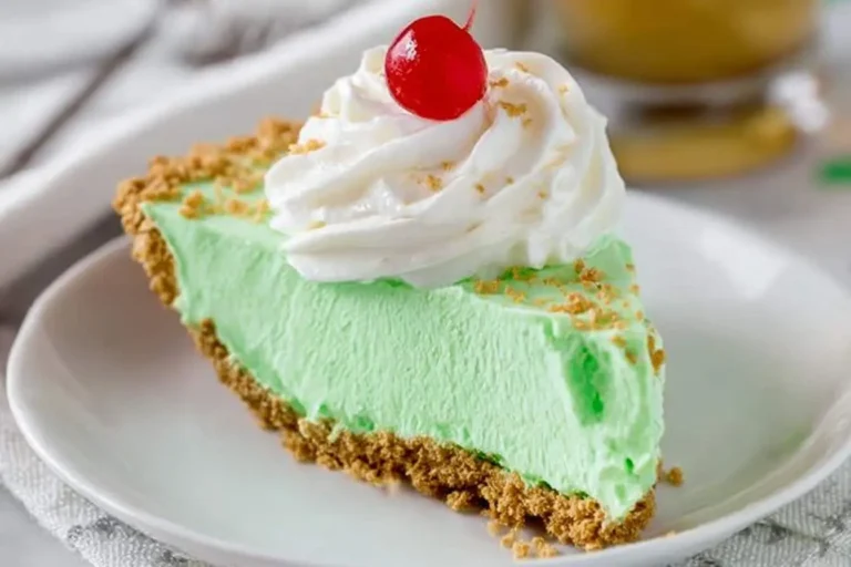 A vibrant slice of shamrock shake pie with a graham cracker crust, topped with fluffy whipped cream and a bright red cherry.