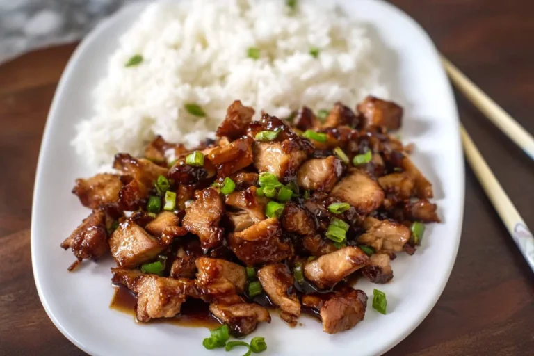 A white plate featuring a generous serving of glossy bourbon chicken garnished with green onions and a side of fluffy white rice.