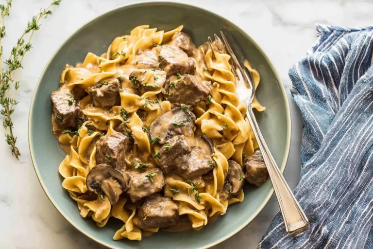 A close-up of a teal bowl filled with creamy crockpot beef stroganoff, featuring wide egg noodles, tender beef, mushrooms, and fresh thyme, with a fork resting in it.