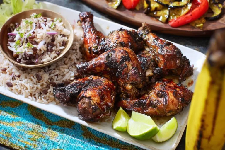 A platter of glossy Jerk Chicken pieces served with traditional rice and peas, garnished with fresh lime wedges.