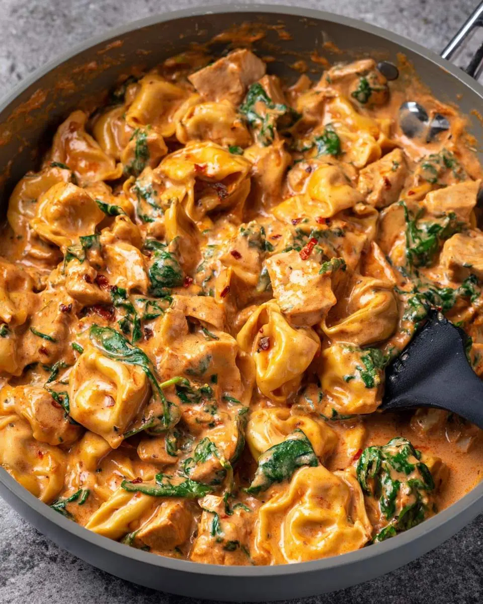 A close-up of creamy Marry Me Tortellini with chicken and spinach in an orange sauce, garnished with red pepper flakes, in a grey pan.