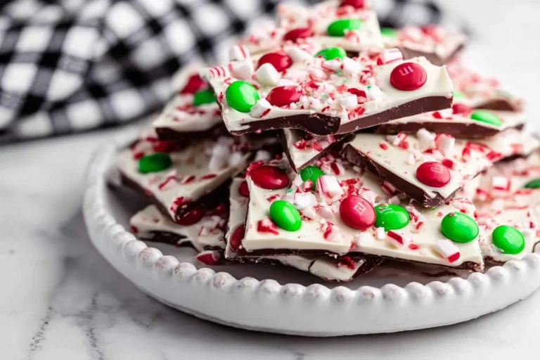 A close-up shot of a white plate piled high with festive peppermint bark, adorned with red and green candies and crushed candy canes.