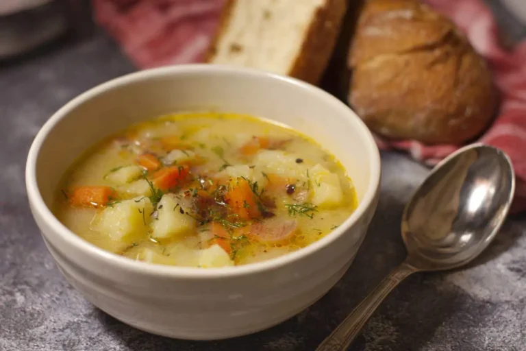 A warm bowl of creamy potato soup, garnished with carrots and fresh dill, served on a stone surface with bread and a spoon.