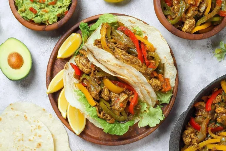 A close-up shot of delicious chicken fajitas on a wooden plate with lettuce and lemon wedges, accompanied by guacamole and extra fajitas in bowls.