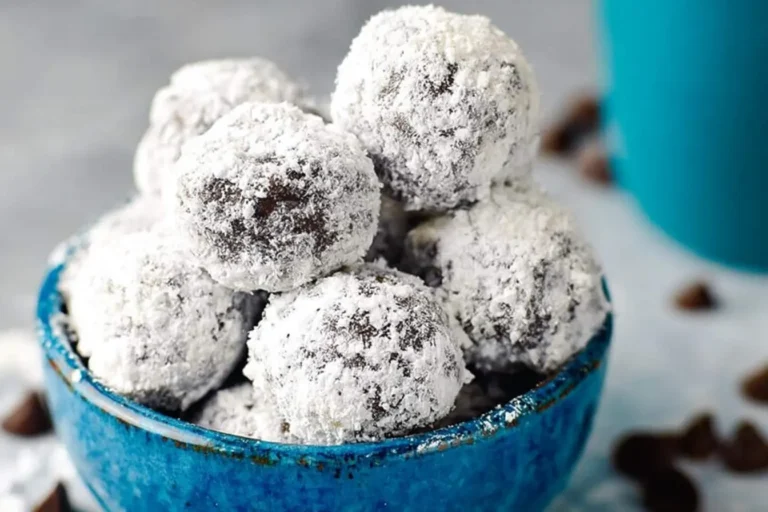 A vibrant blue bowl overflows with a pile of dark chocolate snowballs generously coated in white powdered sugar, with chocolate chips scattered nearby.
