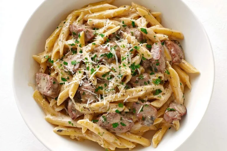 A close-up shot of a white bowl filled with rich and savory creamy steak pasta, garnished with fresh parsley and grated Parmesan cheese.