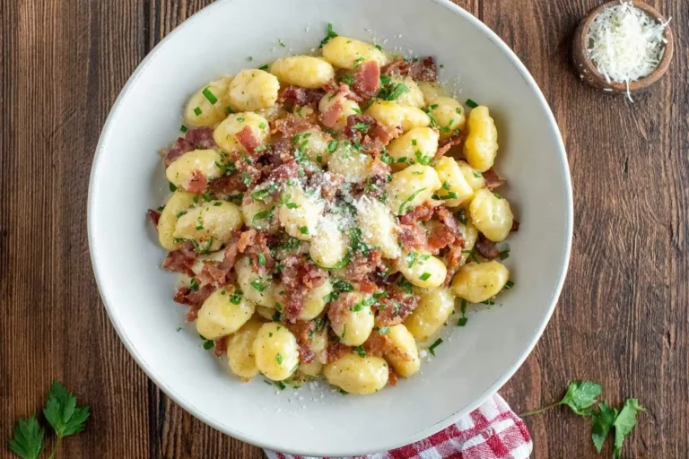 A top-down view of a white bowl filled with creamy Gnocchi Carbonara, topped with crispy bacon, fresh parsley, and grated Parmesan cheese on a wooden table.