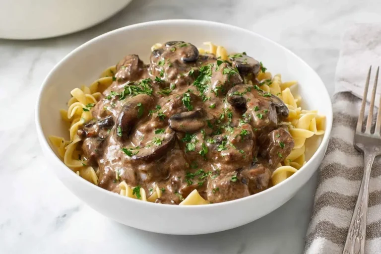 A white bowl of rich Ground Beef Stroganoff with mushrooms and egg noodles, garnished with parsley, on a marble surface.