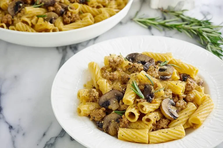 A close-up shot of a white plate filled with creamy ground turkey pasta, sautéed mushrooms, and fresh rosemary sprigs.