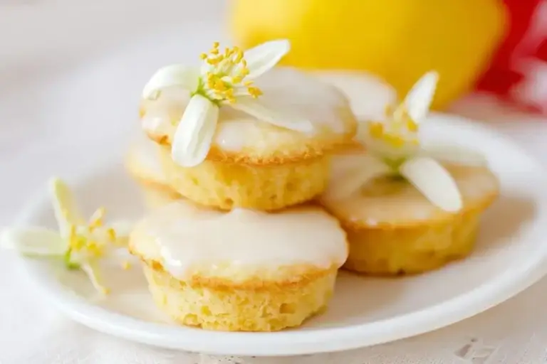 Small lemon cakes with white icing and delicate lemon blossoms on a white plate, with a blurred lemon in the background.