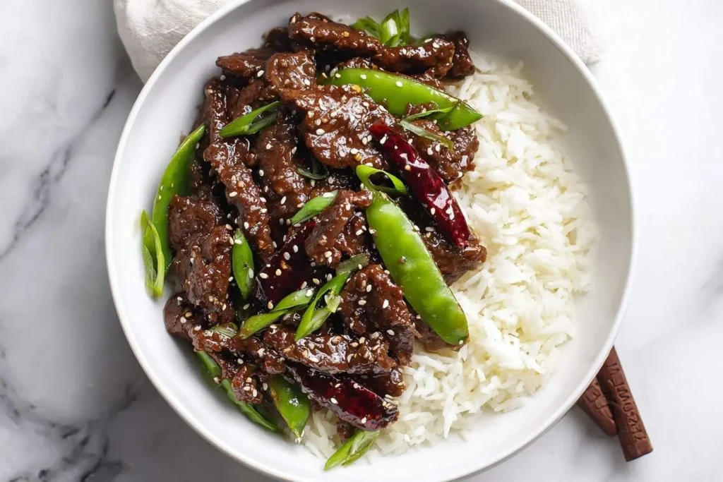 A white bowl holds delicious Mongolian Beef with white rice, green snap peas, and red chilies, sprinkled with sesame seeds.