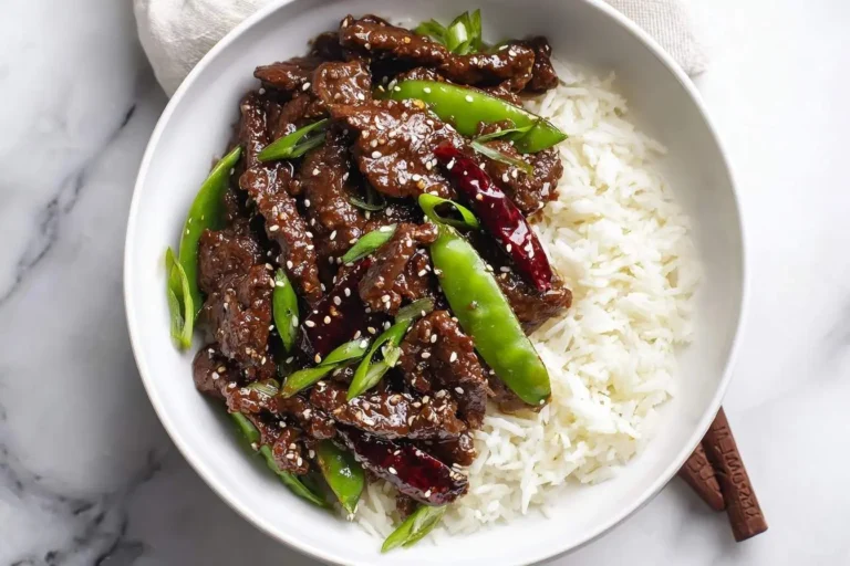 A white bowl holds delicious Mongolian Beef with white rice, green snap peas, and red chilies, sprinkled with sesame seeds.