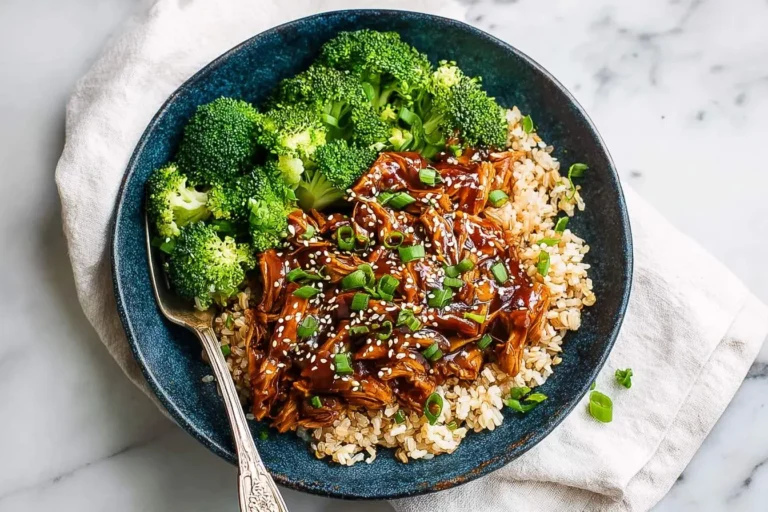 A blue bowl with slow cooker honey garlic chicken, brown rice, and steamed broccoli, garnished with sesame seeds and green onions.