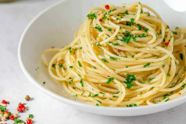 A close-up of a white bowl filled with classic Spaghetti Aglio e Olio, garnished with fresh parsley and red chili flakes.