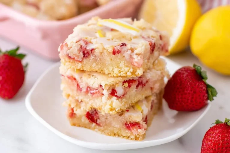 A stack of three strawberry lemon blondies with a white glaze and lemon zest on a white plate, with fresh strawberries and lemons in the background.