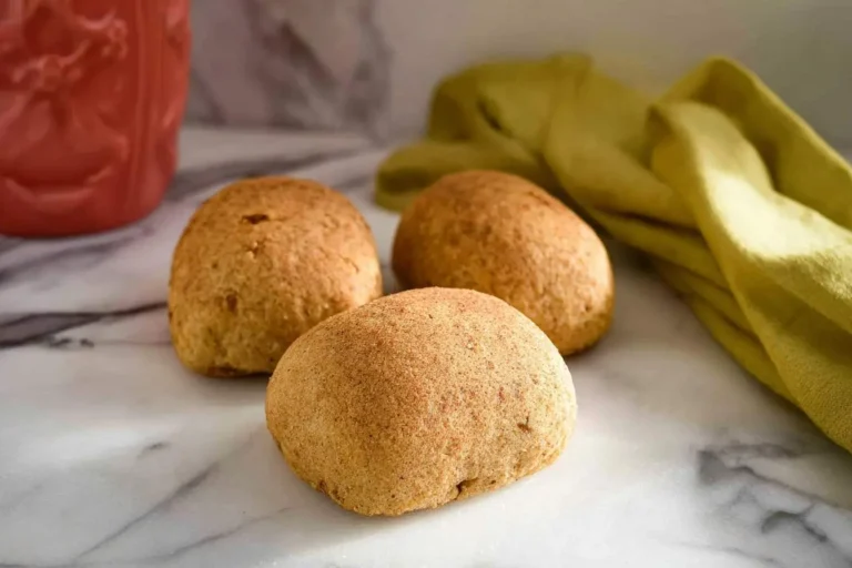 Three golden-brown Almond Flour Rolls displayed on a white marble countertop with a red pitcher and yellow cloth in the background.