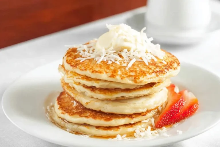A stack of golden coconut cream pancakes topped with shredded coconut and a dollop of butter, served with a fresh strawberry slice.