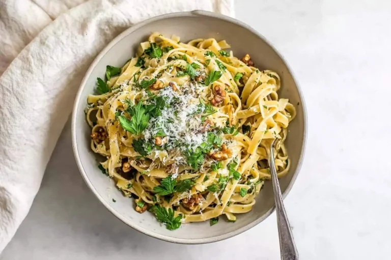 A top-down view of a light grey bowl filled with creamy Fettuccine with Walnuts and Parsley, garnished with grated cheese and a fork, beside a white napkin.