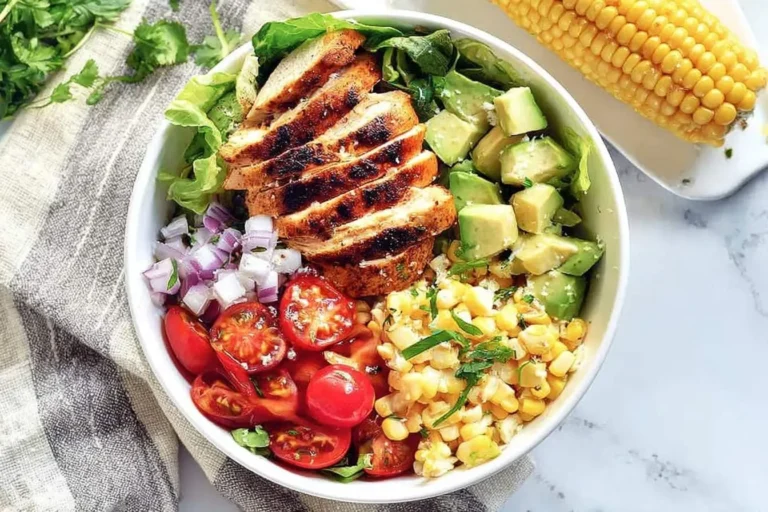 A colorful grilled chicken salad bowl with sliced chicken, avocado, corn, cherry tomatoes, and red onion on a marble countertop.
