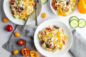 Three vibrant bowls of Mediterranean orzo salad with feta, olives, colorful bell peppers, and fresh vegetables on a light background.