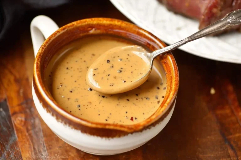 A spoonful of creamy, speckled peppercorn sauce is lifted from a rustic ceramic bowl on a wooden table, ready to serve with steak.