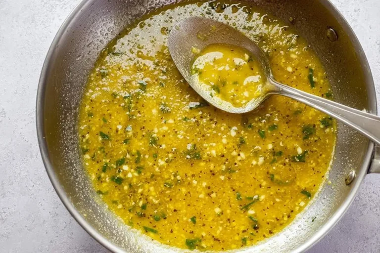 A silver spoon scoops a rich, yellow Spicy Garlic Butter Sauce with visible minced garlic and green herbs in a metal pan.