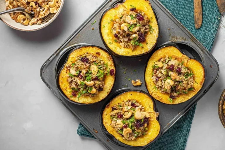 Four golden Stuffed Acorn Squash halves, filled with grains, cranberries, and cashews, baked in a dark metal pan.