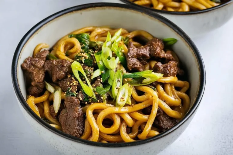 A close-up of a bowl filled with savory beef black pepper udon noodles, garnished with fresh green onions and sesame seeds.