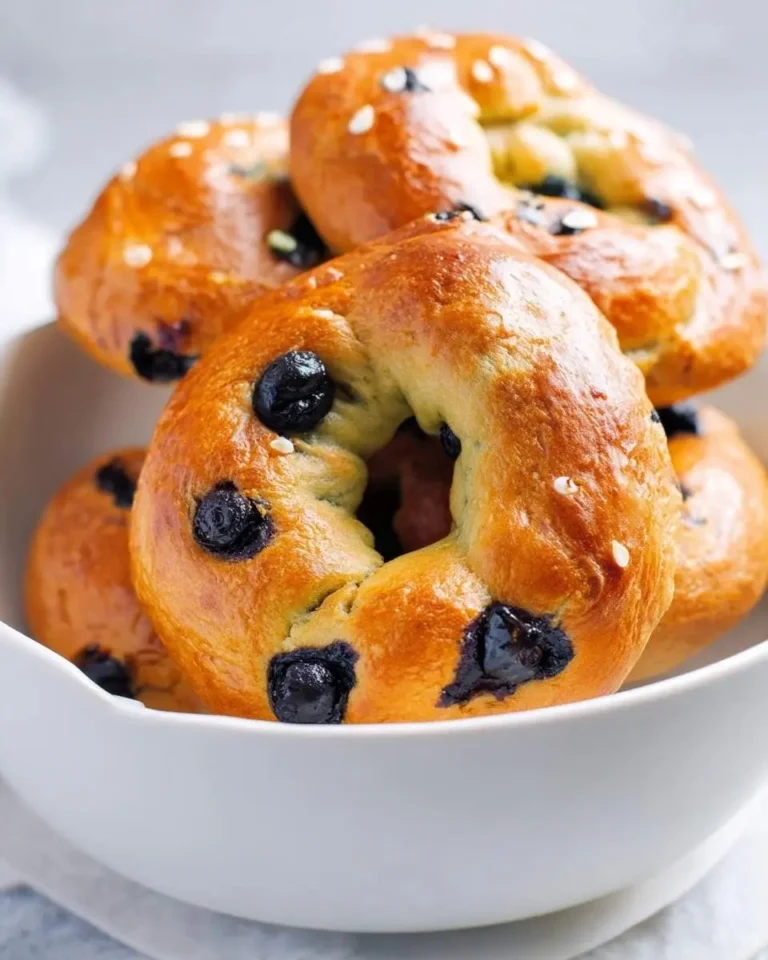A close-up view of several fresh, golden-brown blueberry bagels, some sprinkled with sesame seeds, stacked in a white bowl.