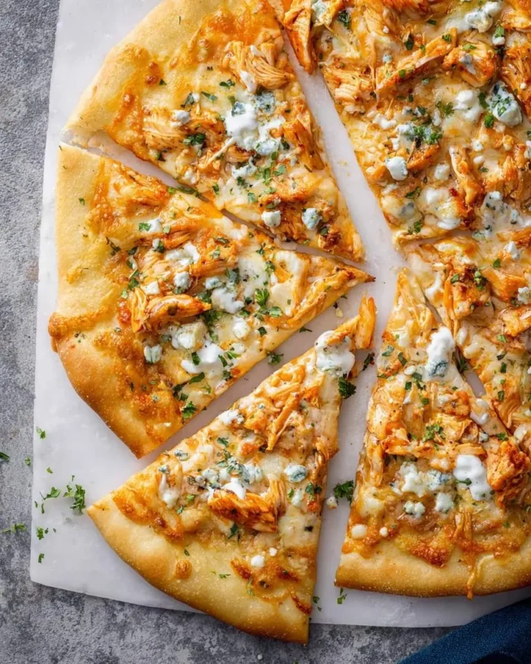 A delicious Buffalo Chicken Pizza, generously topped with shredded chicken, blue cheese, and fresh herbs, cut into slices on a white surface.