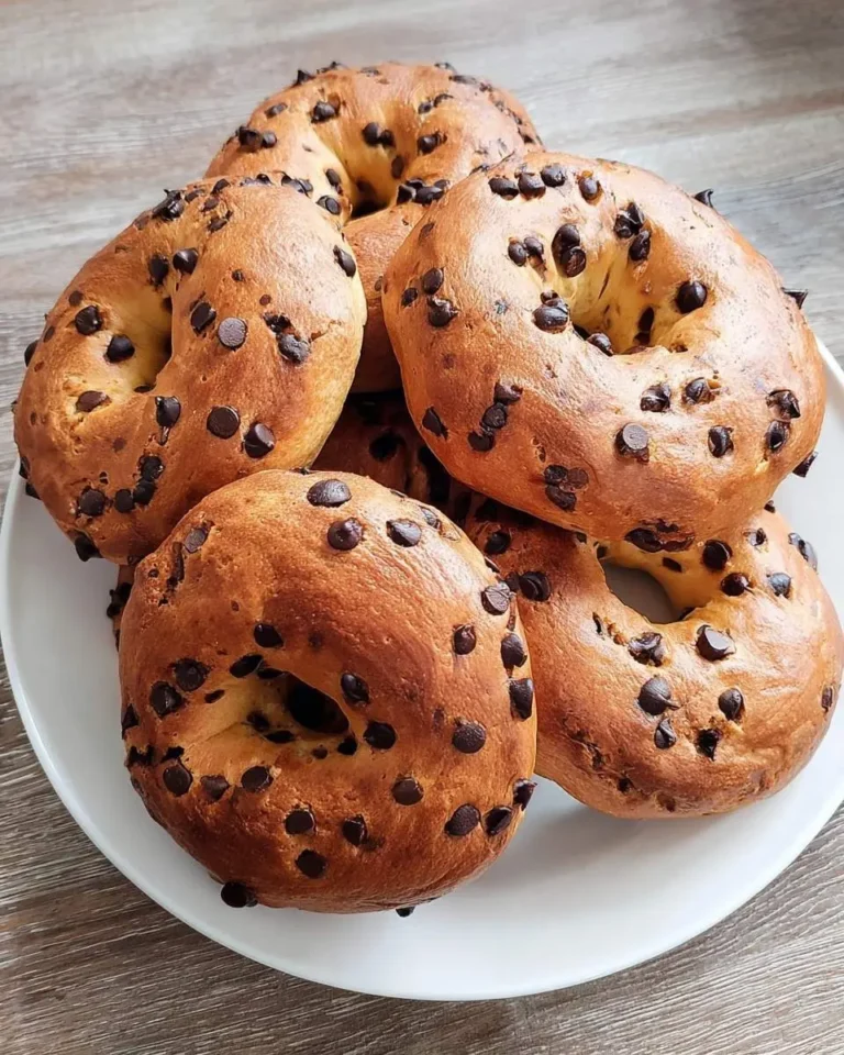 A white plate filled with golden-brown chocolate chip bagels, generously sprinkled with chocolate chips, on a wooden surface.