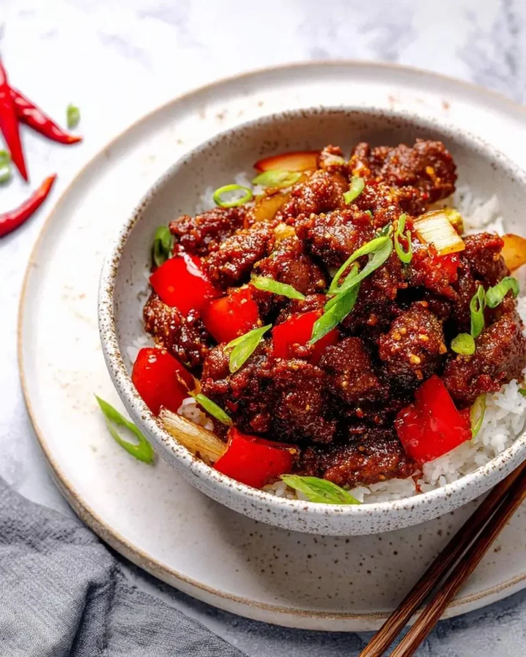 A bowl of savory Crispy Beijing Beef served over white rice, garnished with red bell peppers and fresh green onions, with chopsticks on the side.