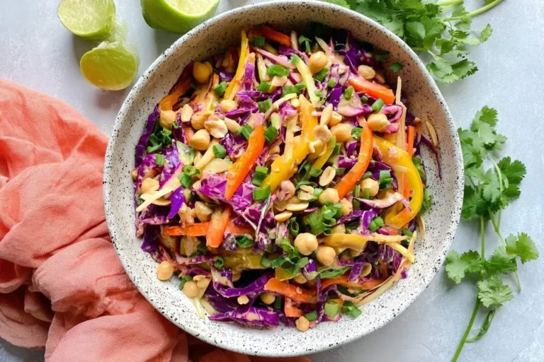A vibrant bowl of Crunchy Thai Chickpea Salad with purple cabbage, colorful bell peppers, chickpeas, peanuts, and green onions, garnished with lime wedges.