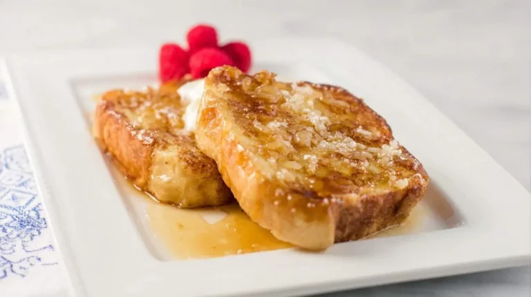 Two slices of golden Crème Brûlée French Toast drizzled with syrup, topped with whipped cream and fresh raspberries on a white plate.