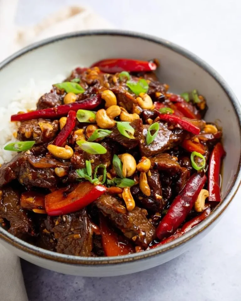 A close-up shot of a bowl of delicious Kung Pao Beef served over white rice, garnished with roasted cashews, vibrant red chili peppers, and fresh green onions.