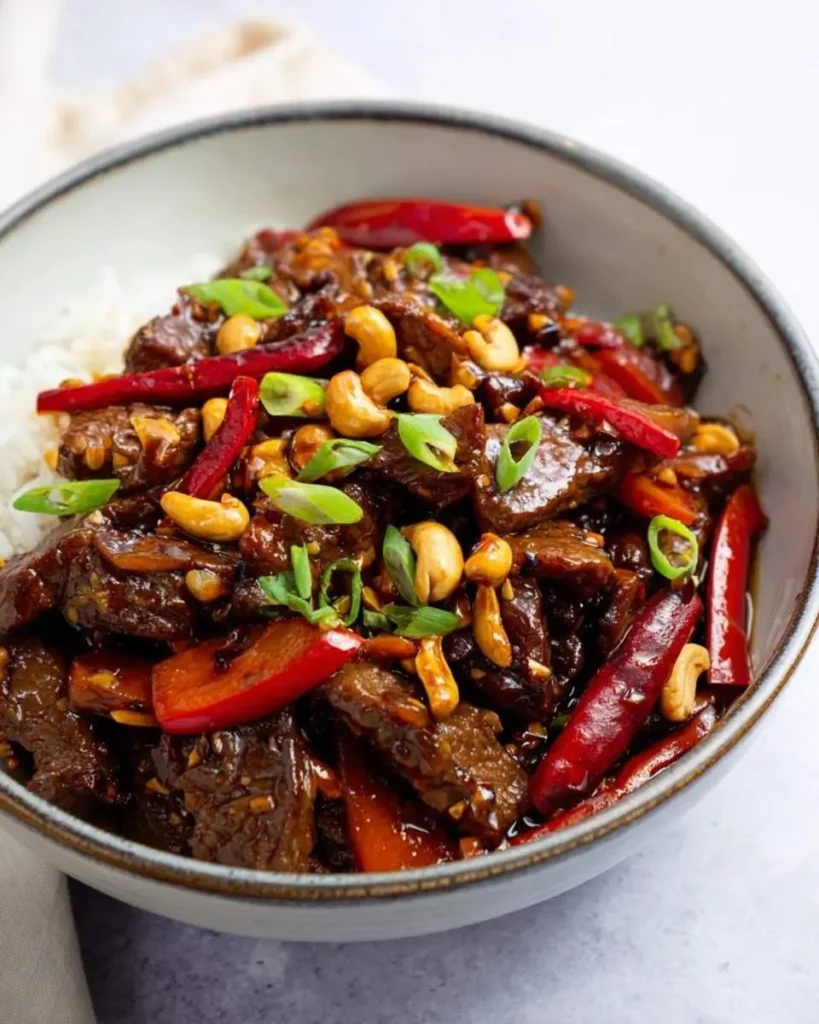 A close-up shot of a bowl of delicious Kung Pao Beef served over white rice, garnished with roasted cashews, vibrant red chili peppers, and fresh green onions.