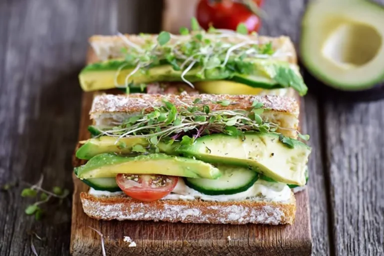 A stacked Loaded Cucumber and Avocado Sandwich on crusty bread with tomato, cucumber, avocado slices, and sprouts, on a wooden board.