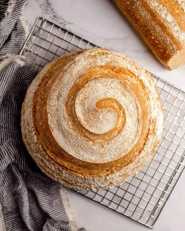 Top-down view of a beautifully scored round No Knead Whole Wheat Bread loaf on a cooling rack, with a baguette in the background.