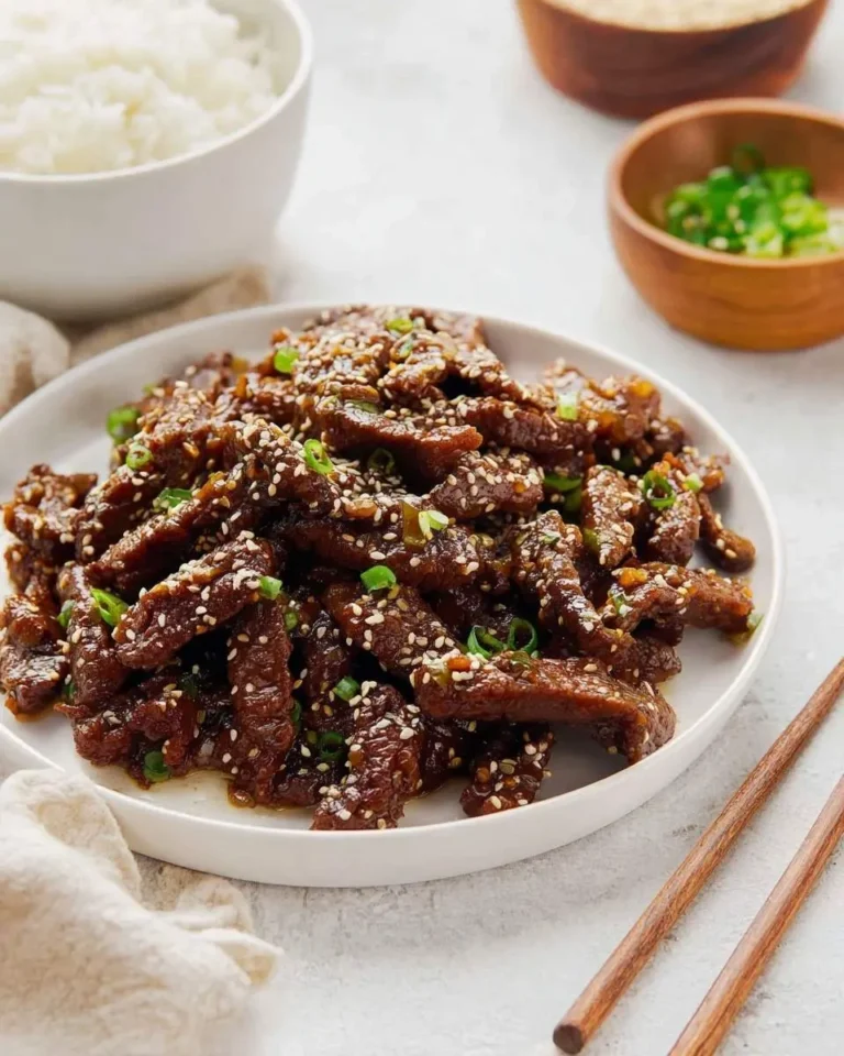 A white plate piled high with glistening Sesame Beef strips, generously sprinkled with sesame seeds and green onions, served alongside a bowl of white rice.