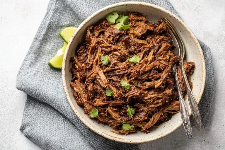 A white bowl brimming with tender Slow Cooker Beef Barbacoa, garnished with fresh cilantro and lime wedges, with two forks.
