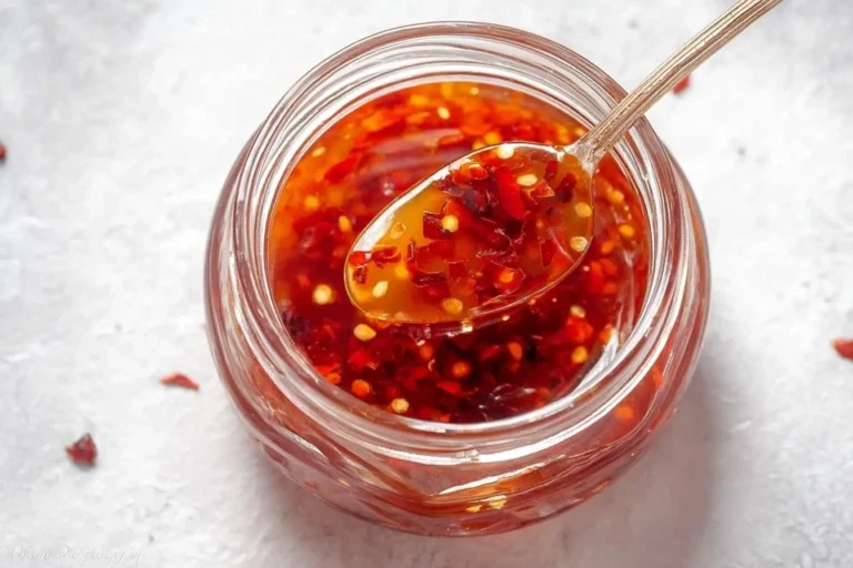 A spoon lifts vibrant red sweet chili sauce, chunky with chili flakes and seeds, from a clear glass jar on a light background.
