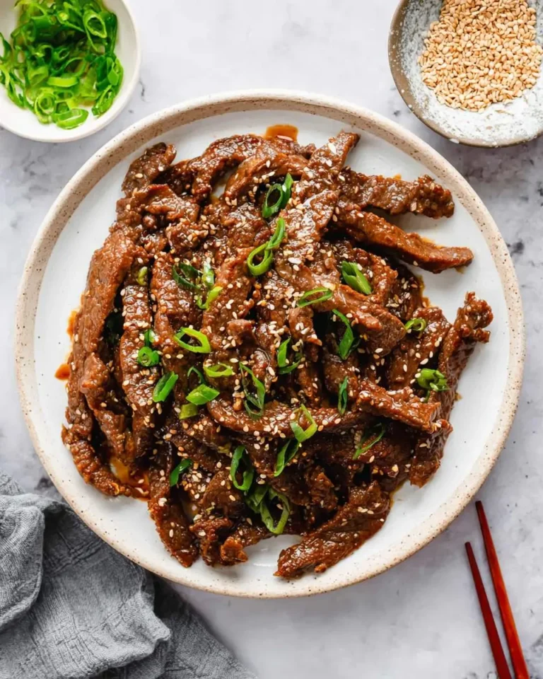 A plate of glistening Beef Bulgogi, garnished with white sesame seeds and fresh green onions, surrounded by garnishes.