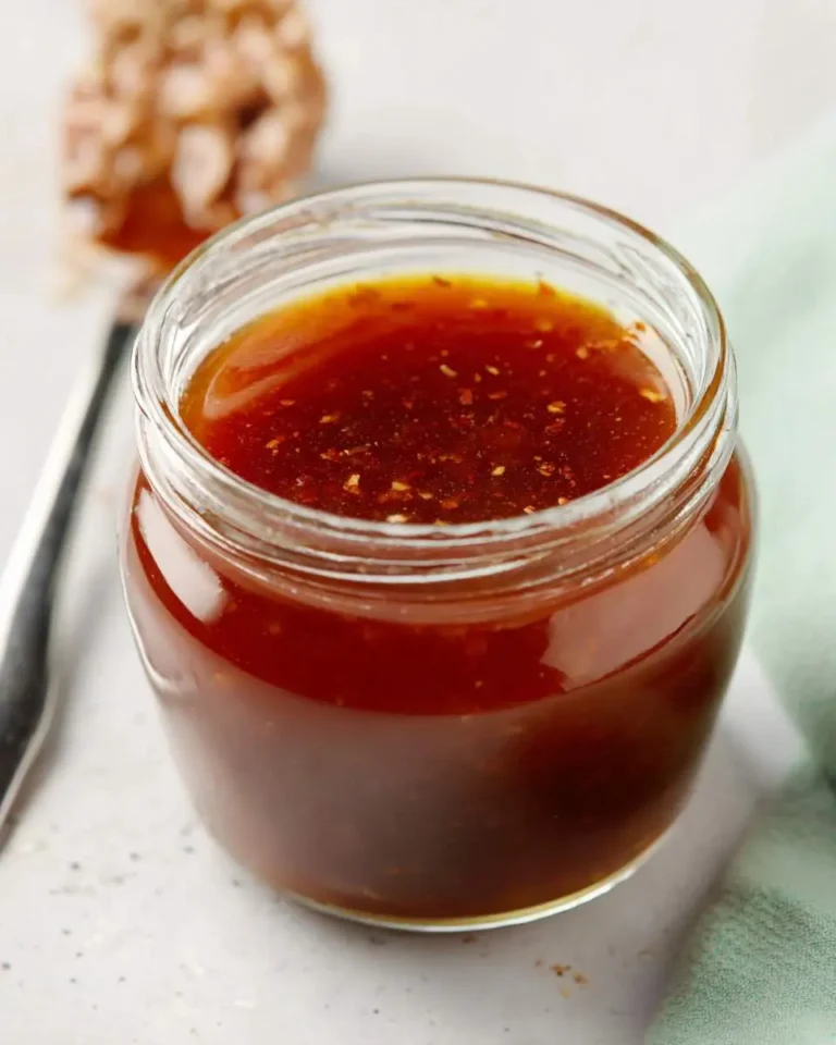 A clear glass jar filled with a reddish-brown Carolina Vinegar BBQ Sauce, showing specks of spices, on a light surface.