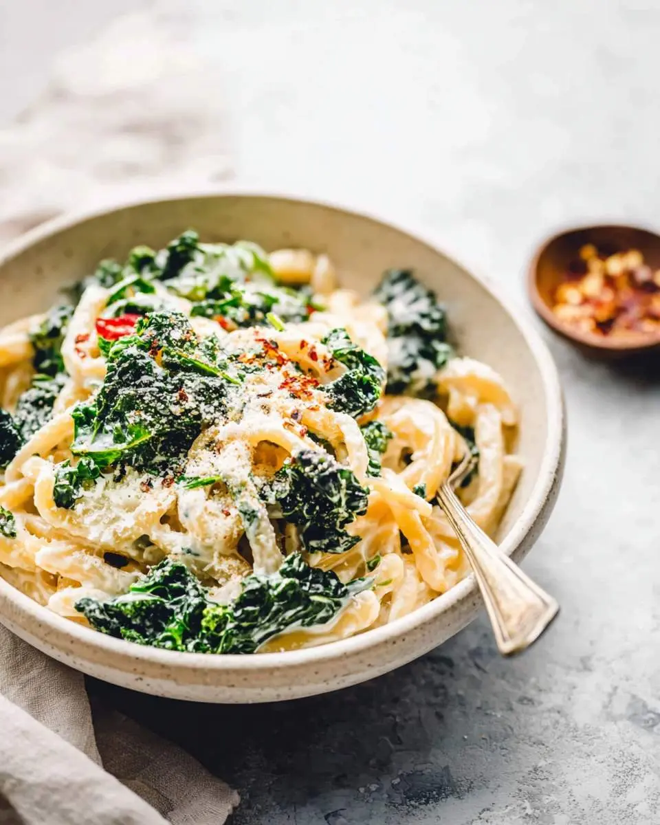 A close-up shot of a bowl filled with creamy kale pasta, garnished with grated cheese and red pepper flakes, with a fork resting inside.