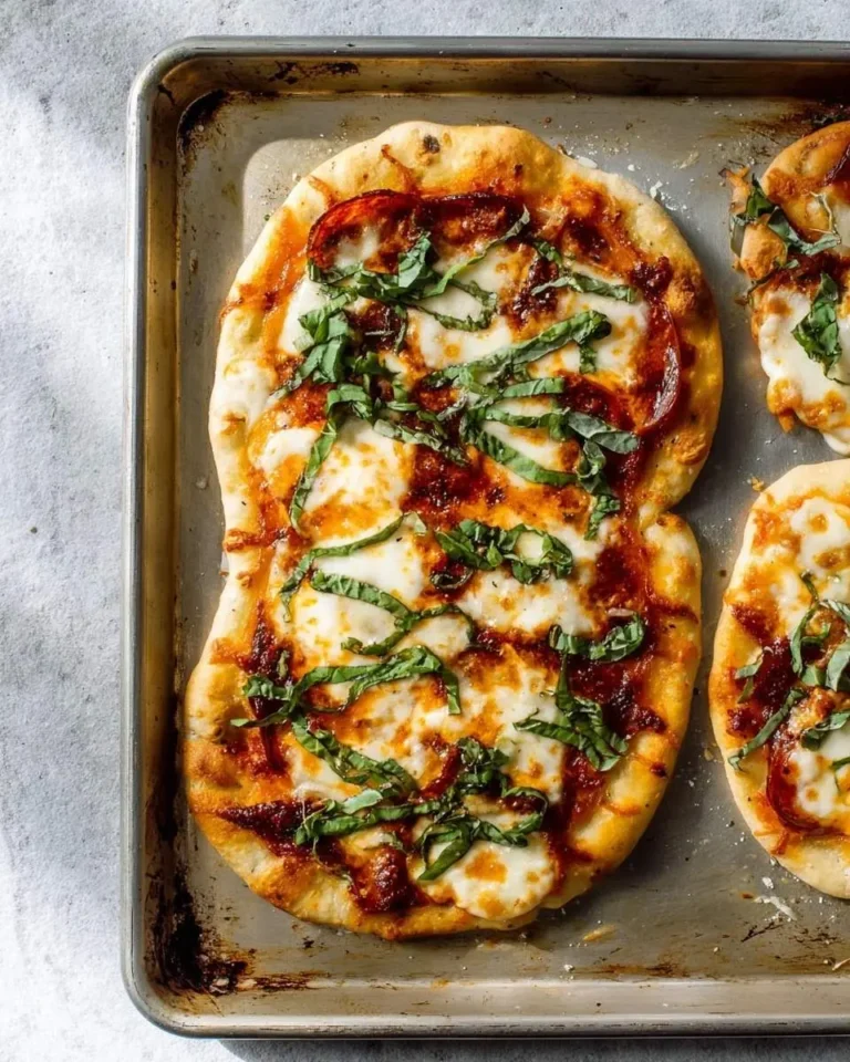 A close-up shot of a large, freshly baked Flatbread Pizza on a metal baking sheet, topped with melted mozzarella, red sauce, and fresh basil leaves. Smaller flatbreads are partially visible.