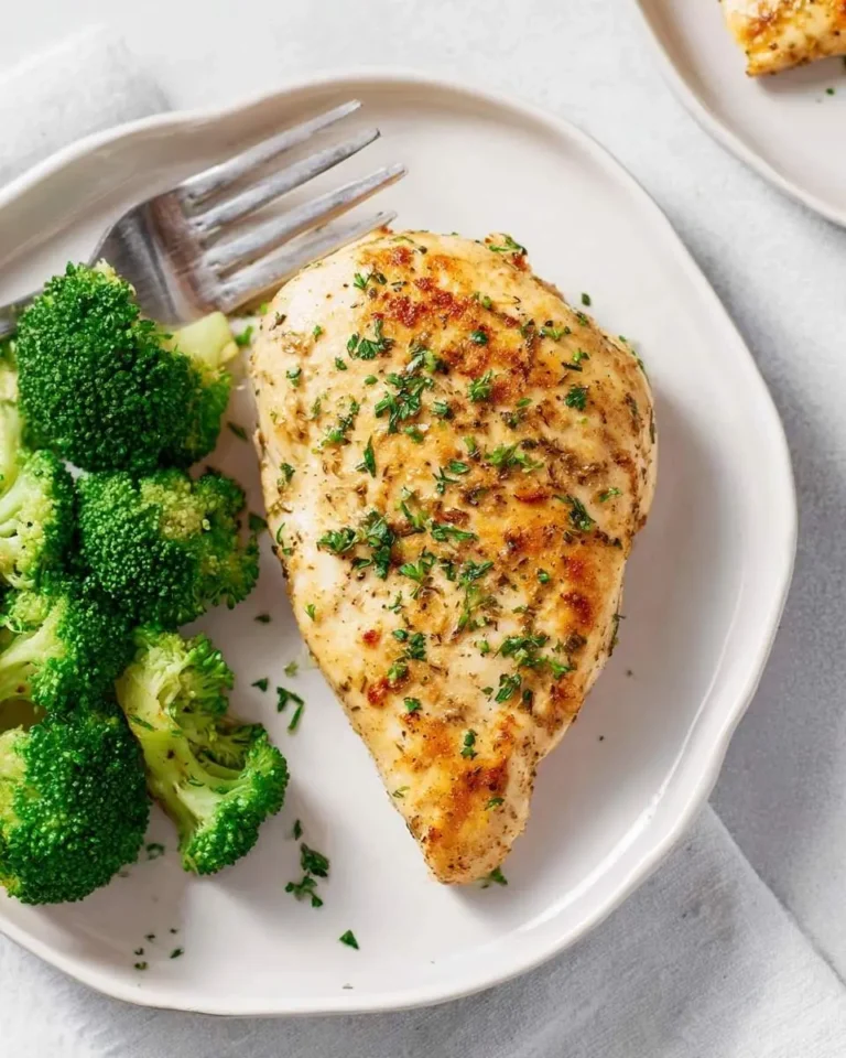 A perfectly cooked Ranch Baked Chicken Breast seasoned with herbs is served alongside fresh green broccoli florets on a white plate with a fork.