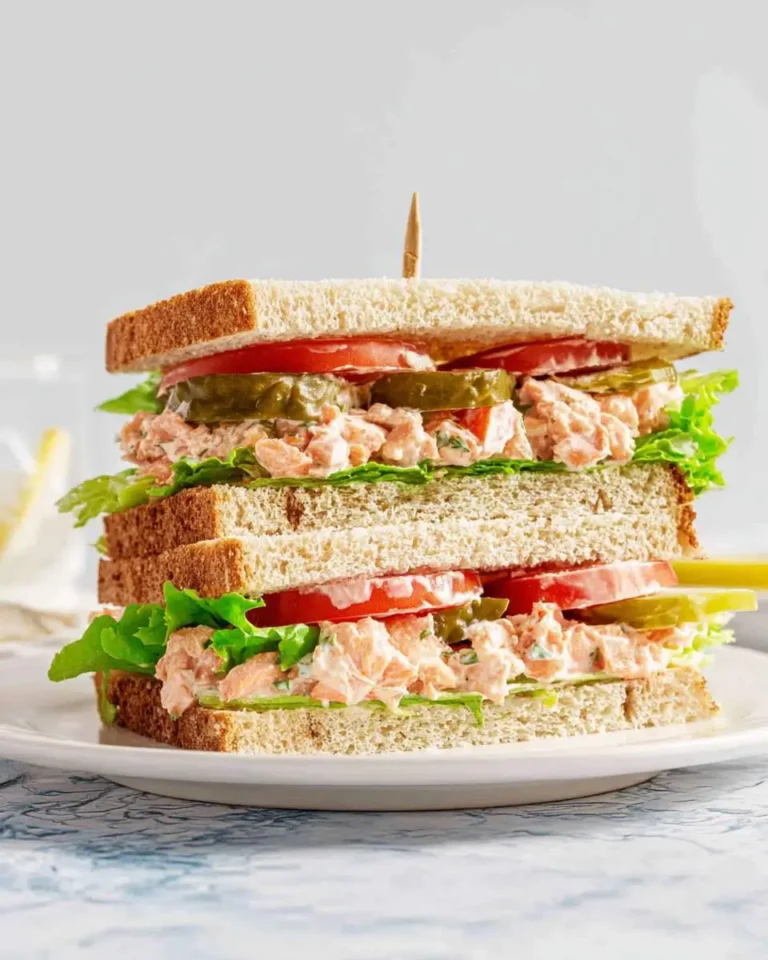 A tall, double-decker Salmon Salad Sandwich with fresh lettuce, tomato slices, and dill pickles, held by a toothpick on a white plate.