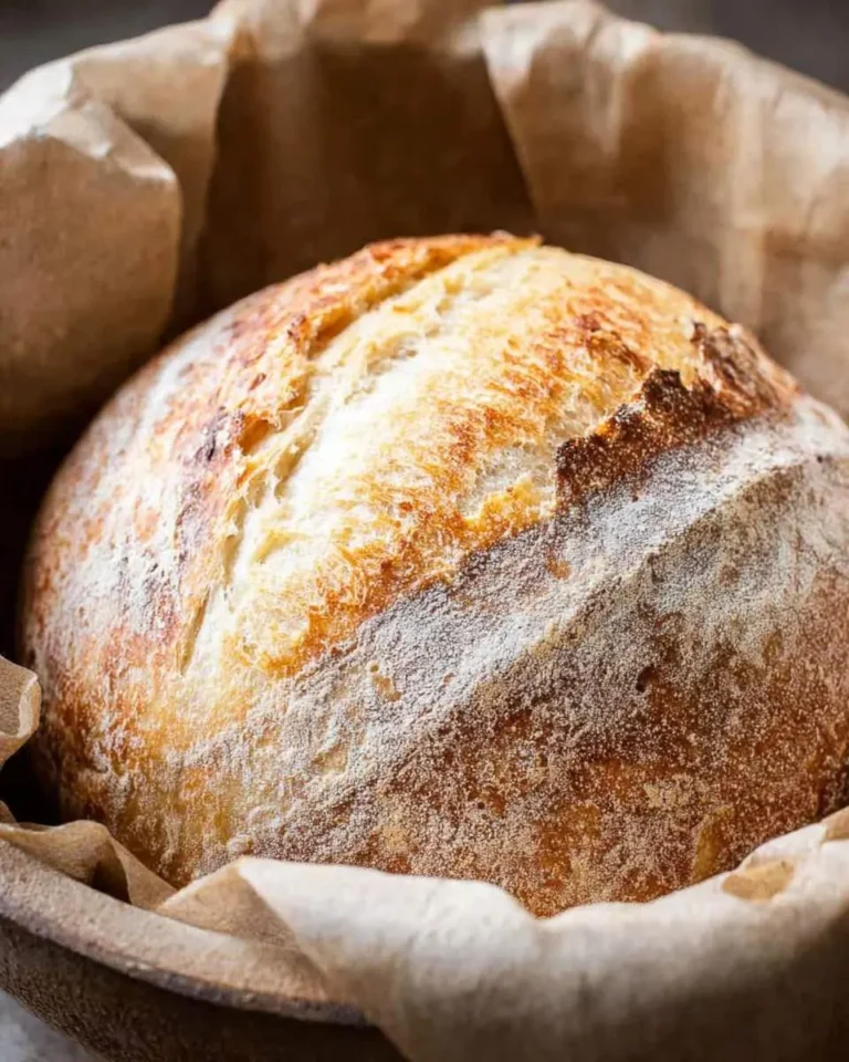 A rustic, golden-crusted loaf of sourdough bread, dusted with flour and resting in a brown paper-lined basket.