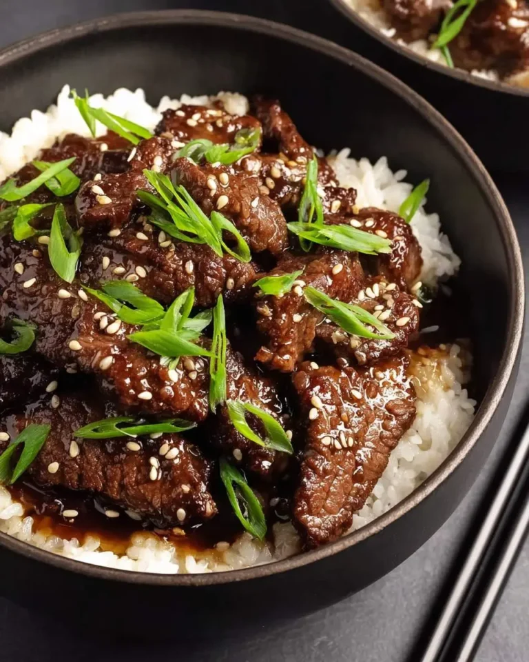 A close-up of a black bowl filled with fluffy white rice topped with glistening teriyaki beef pieces, garnished with fresh green onions and sesame seeds, with chopsticks visible on the side.