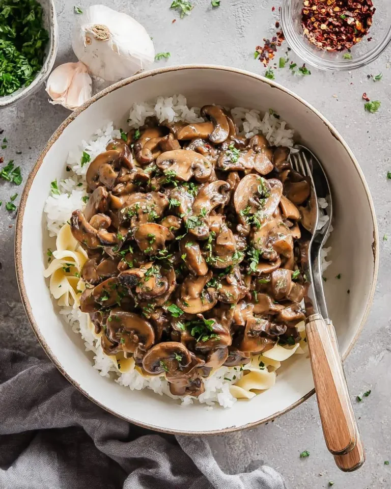 A bowl of creamy Vegan Mushroom Stroganoff served over white rice and egg noodles, garnished with fresh parsley, alongside garlic.